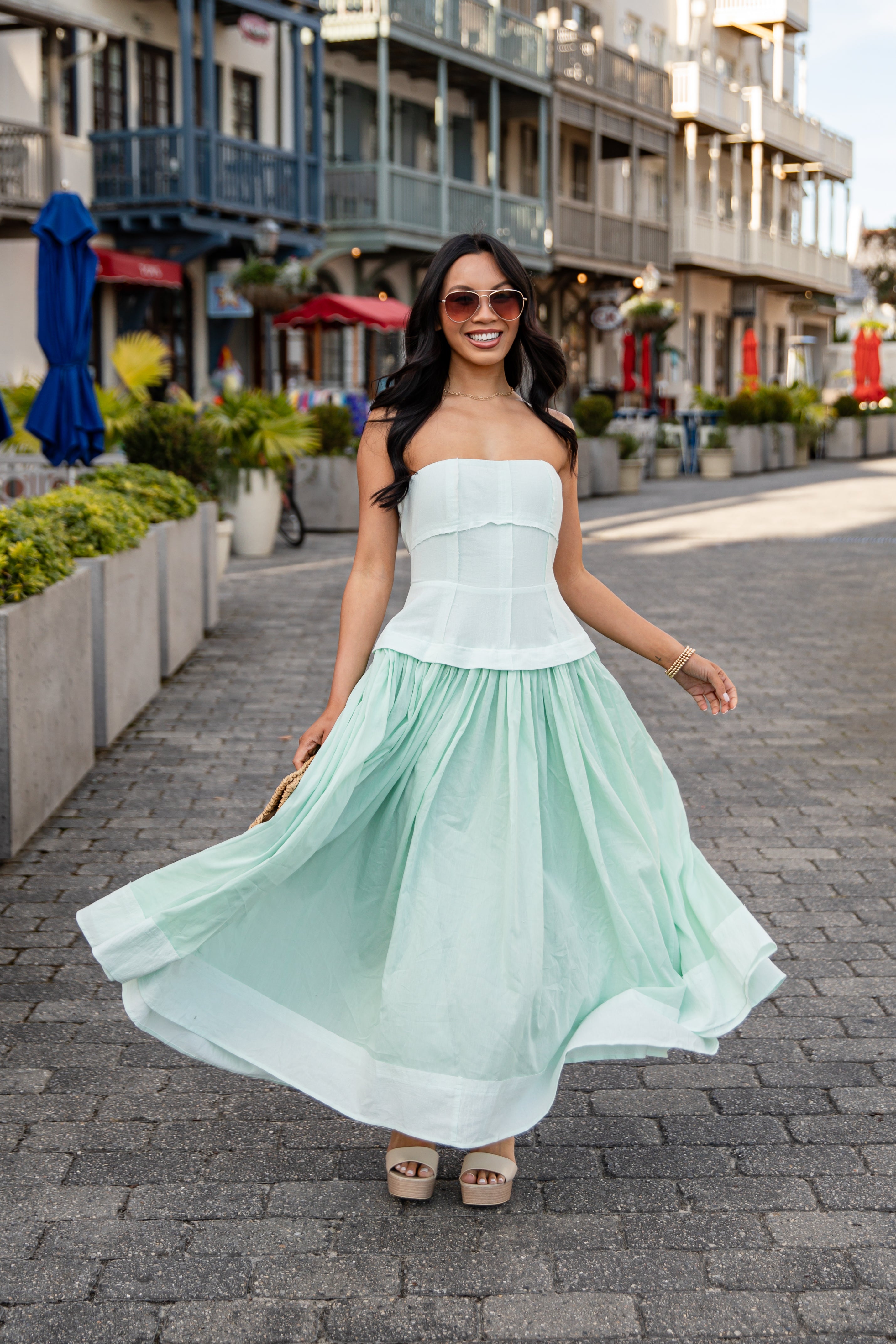 Woman in a light green dress walking on a street with buildings and plants in the background