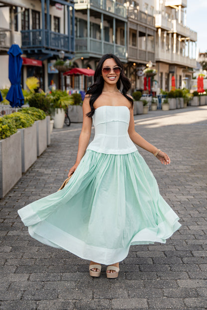 Woman in a light green dress walking on a street with buildings and plants in the background