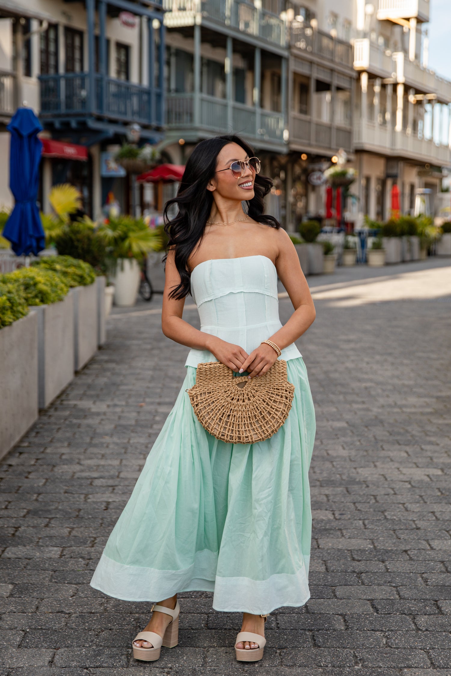 Woman in a strapless white top and light green skirt with a woven handbag on a street.