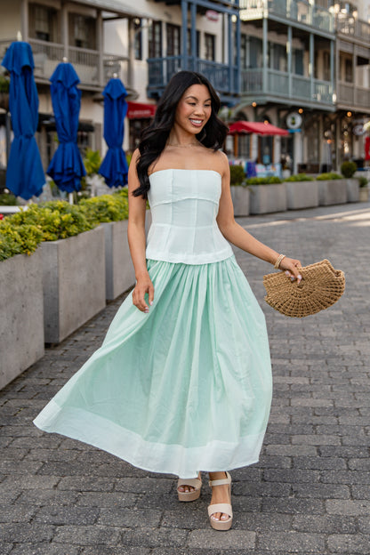 Woman in a light green dress standing on a street with blue umbrellas and buildings in the background.