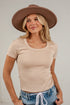 A woman wearing a beige short-sleeve scoop-neck t-shirt, blue jeans with a white drawstring, and a brown wide-brim hat, posing against a white paneled wall.