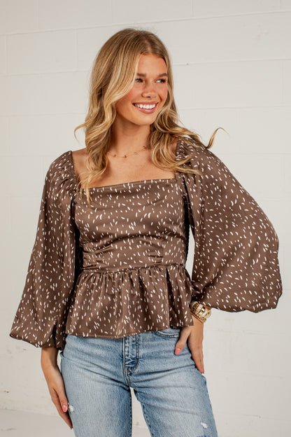 A woman wearing a brown off-the-shoulder blouse with a white speckled pattern, square neckline, and voluminous bell sleeves, paired with light blue jeans, standing against a white brick wall.