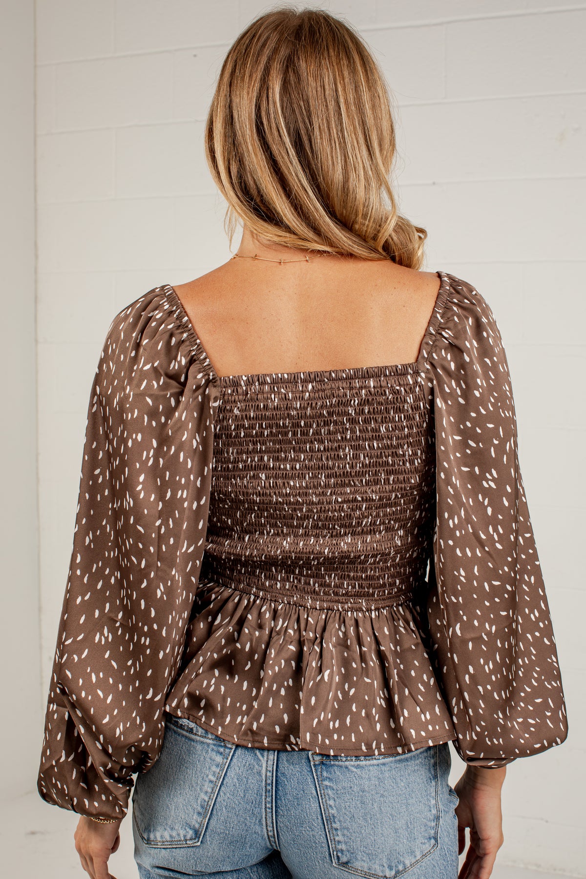 A woman viewed from the back wearing a brown smocked blouse with a white speckled pattern, long puff sleeves, and a square neckline, paired with light blue jeans against a white paneled wall.