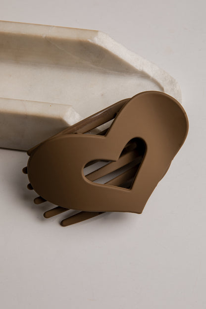 A heart-shaped brown hair claw clip with a cutout heart design, resting on a light gray surface next to a white marble prop.