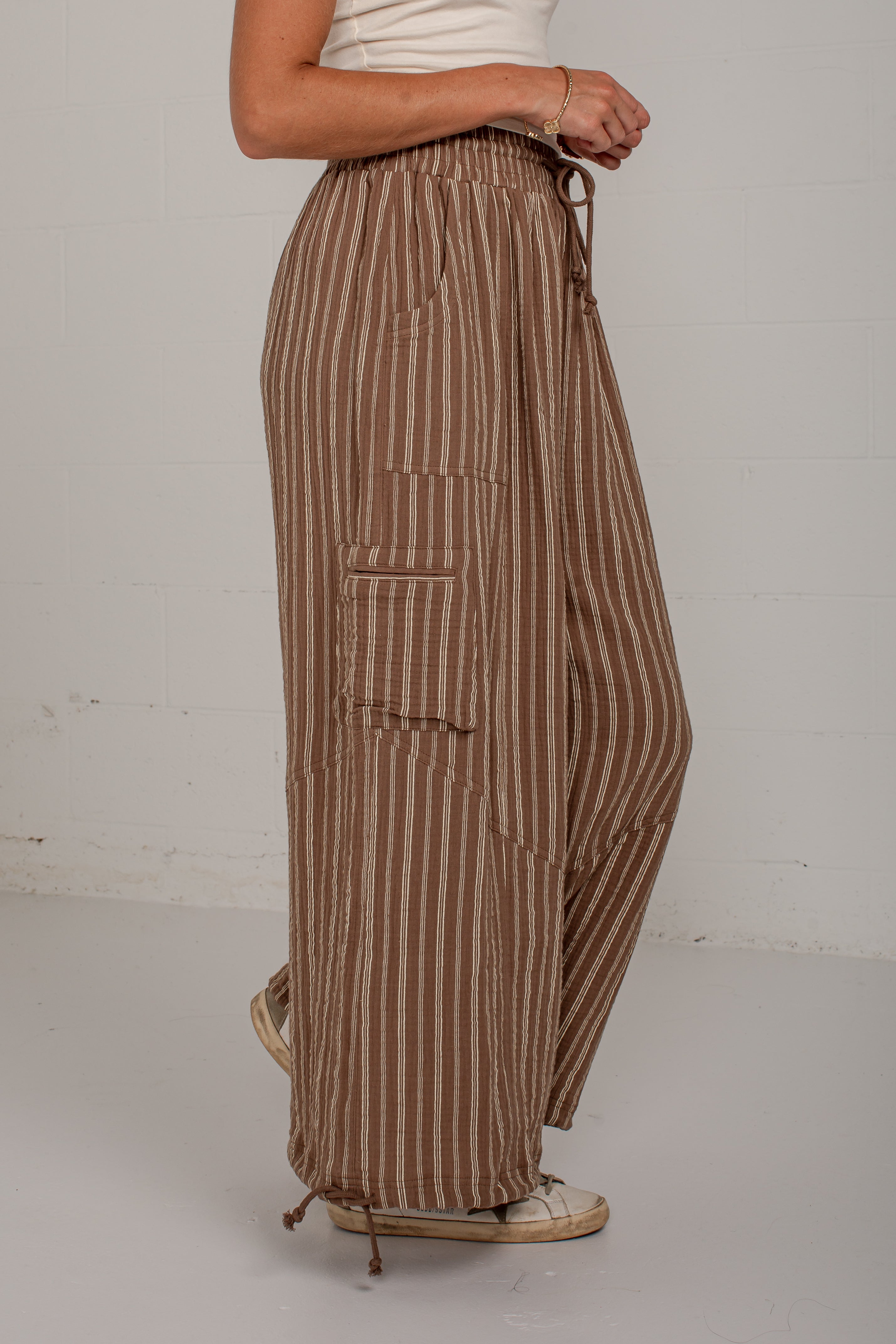 A person wearing loose brown and tan vertically striped wide-leg pants with a drawstring waist and side cargo pocket, shown from the waist down against a plain light gray wall.