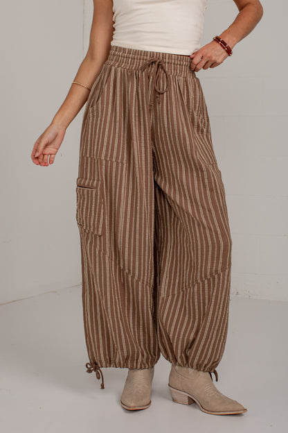 A person wearing loose brown and beige vertically striped wide-leg pants with a drawstring waist, side cargo pockets, and ankle ties, paired with a white top and beige ankle boots against a plain light gray studio background.
