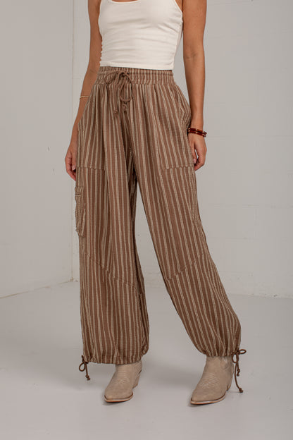 A model wearing brown and beige vertically striped wide-leg pants with a drawstring waist, cargo pockets, and ankle ties, styled with a white tank top and beige ankle boots.