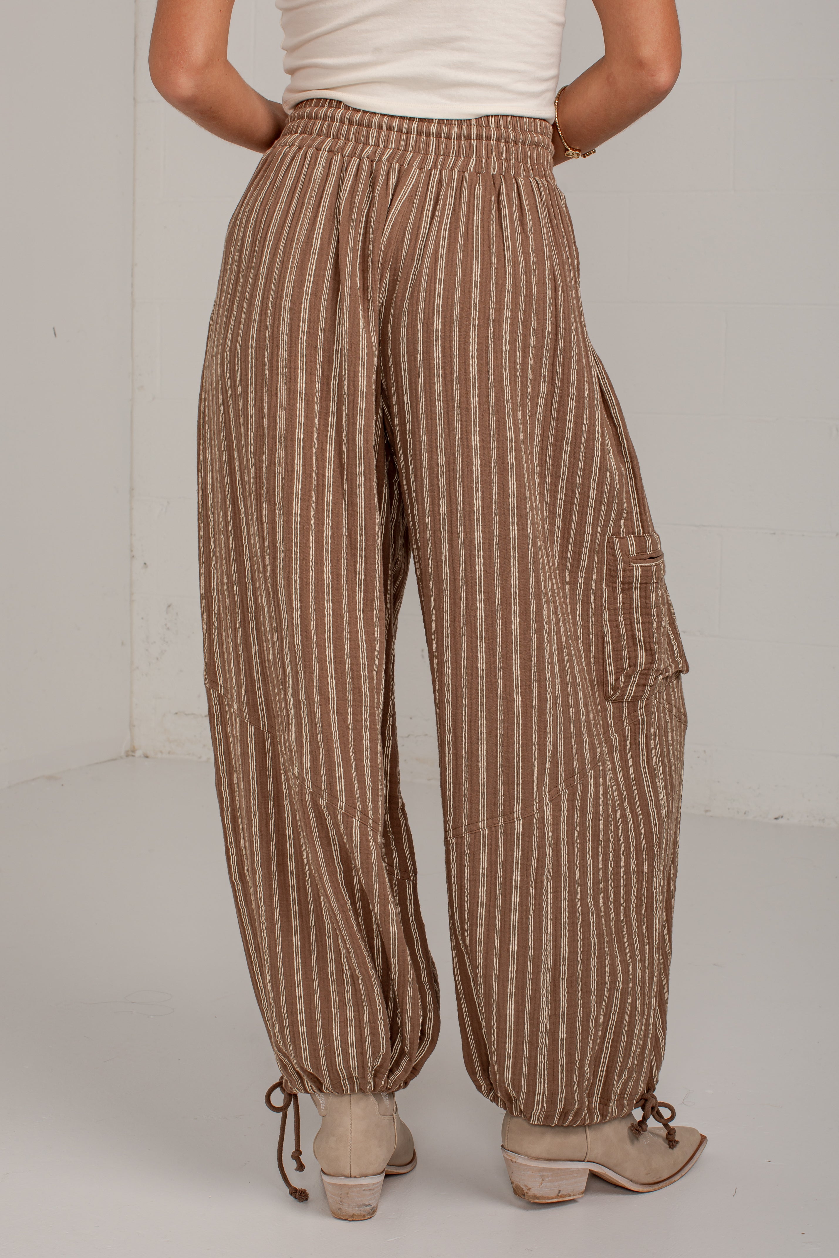 A person shown from the back wearing loose brown and beige vertically striped cargo pants with drawstring hems and light beige heeled sandals, standing against a plain light gray wall.
