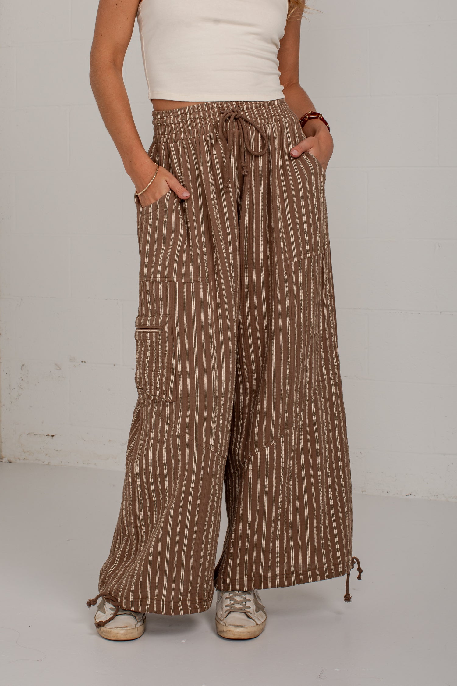 A person wearing brown and beige vertically striped wide-leg pants with side cargo pockets and a drawstring waist, paired with a white ribbed top and beige sneakers, standing against a white wall.