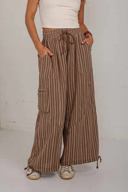 A person wearing brown and beige vertically striped wide-leg pants with side cargo pockets and a drawstring waist, paired with a white ribbed top and beige sneakers, standing against a white wall.