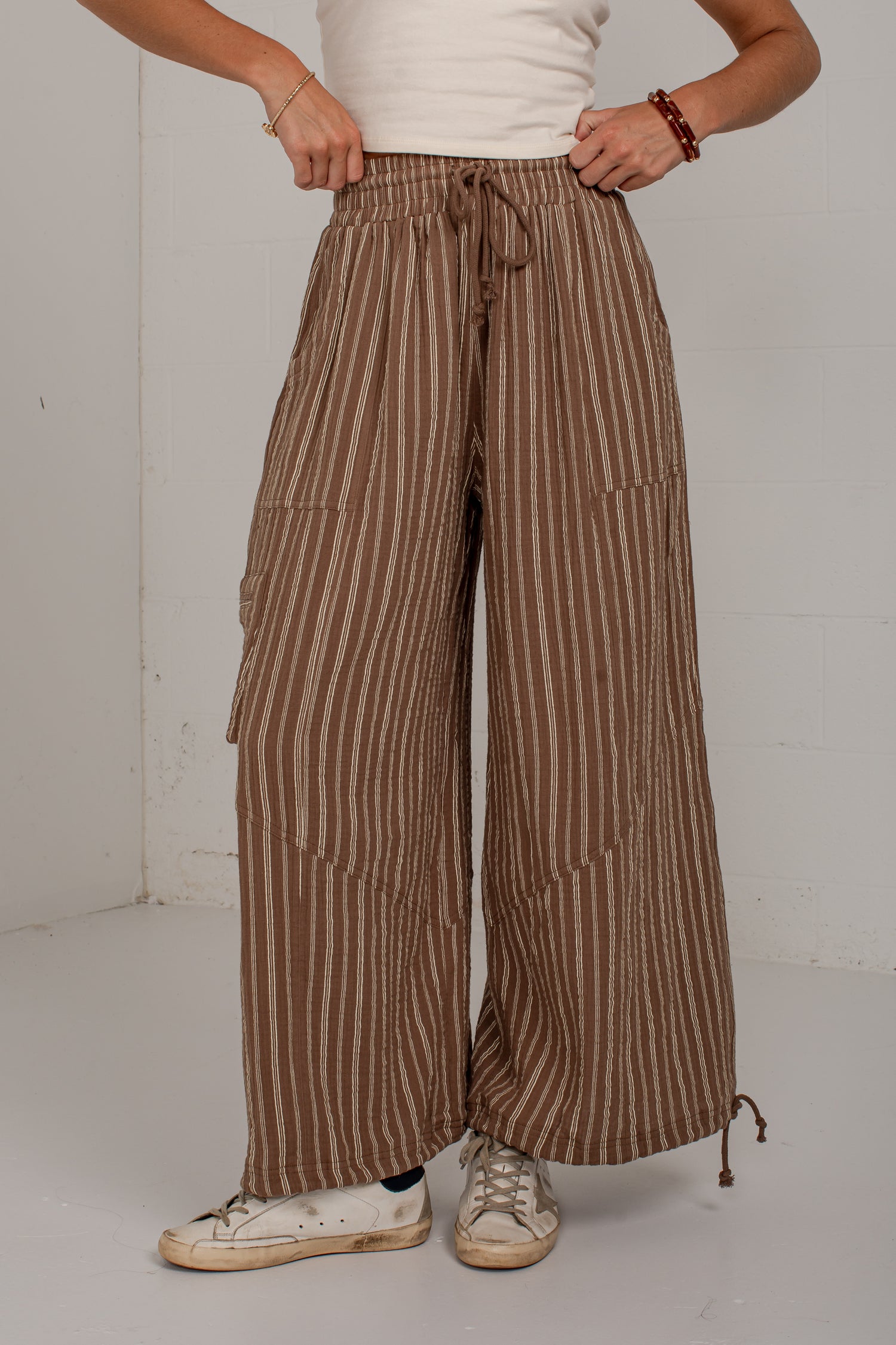 A person wearing brown and beige vertically striped wide-leg pants with a drawstring waist, paired with a white top and white sneakers, standing against a plain light gray wall.