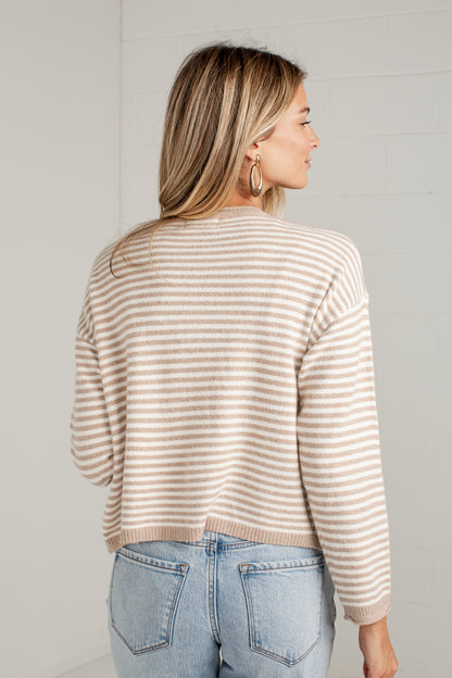 A woman shown from the back wearing a beige and white horizontally striped knit sweater and light blue jeans, standing against a white brick wall.