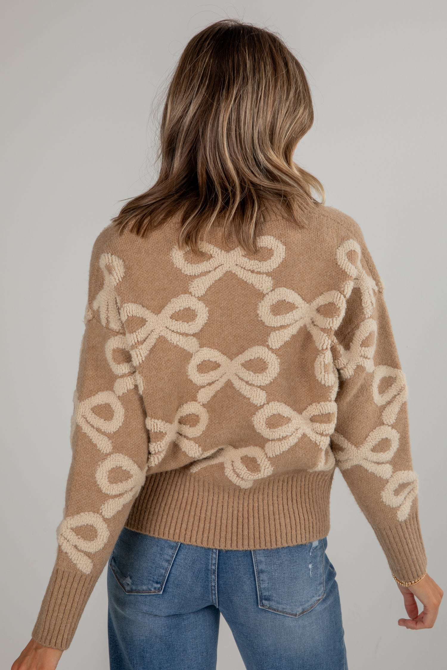Person wearing a brown sweater with bow pattern and blue jeans against a plain background