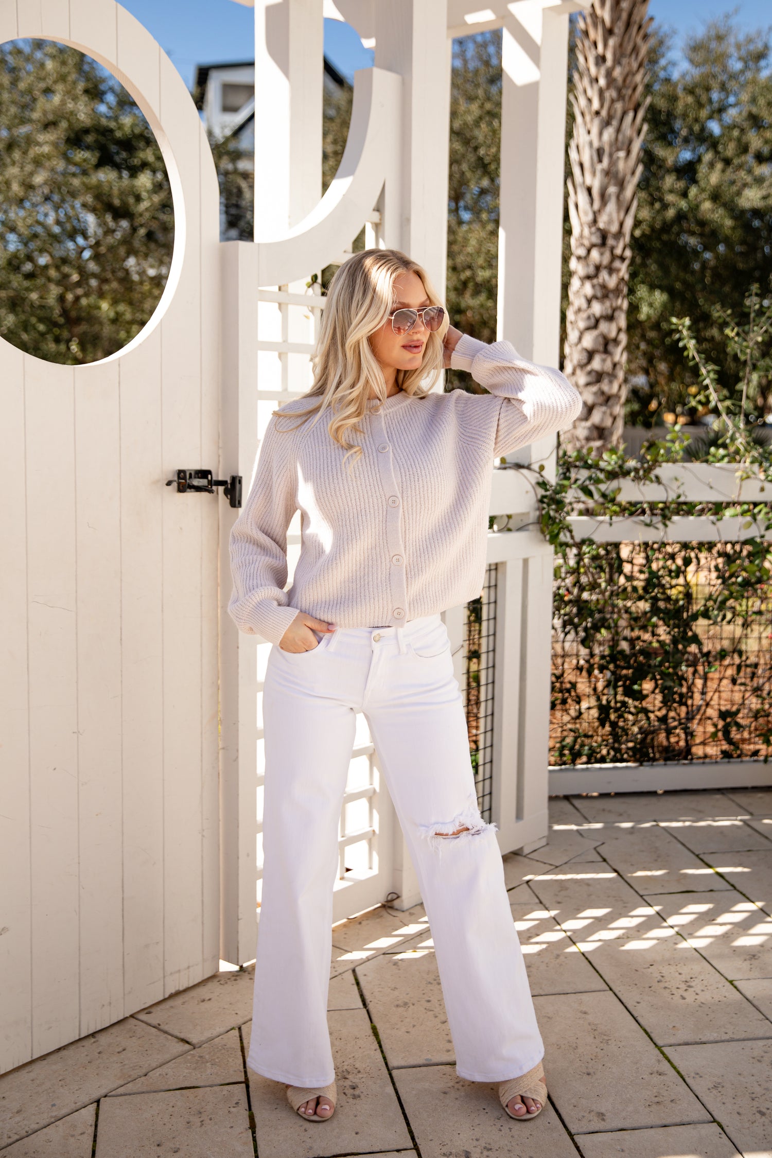 Woman in a light gray sweater and white pants standing in front of a white gazebo.
