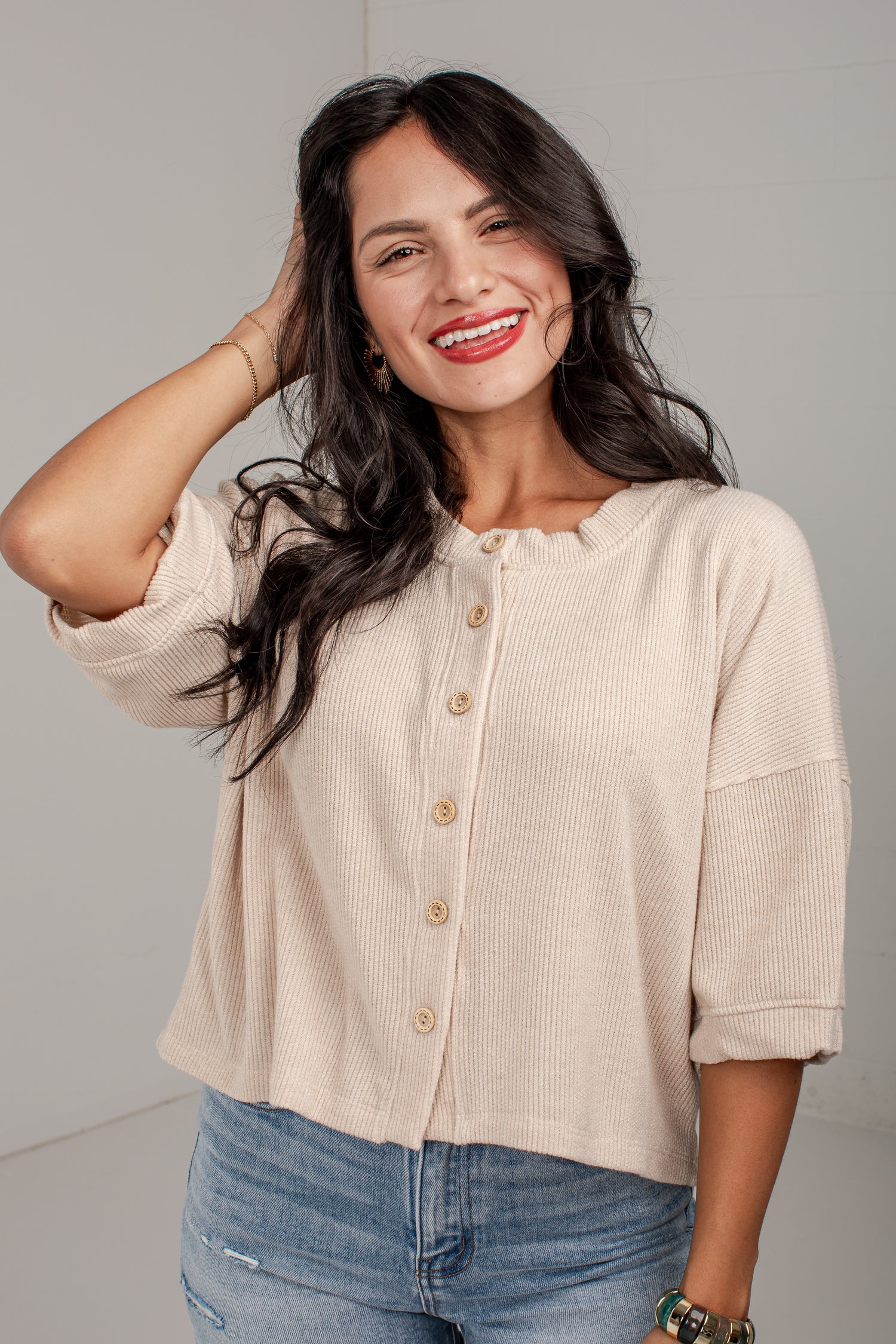A woman with long dark hair smiling while wearing a beige ribbed knit button-up top with rolled sleeves and blue jeans, set against a plain light gray background.