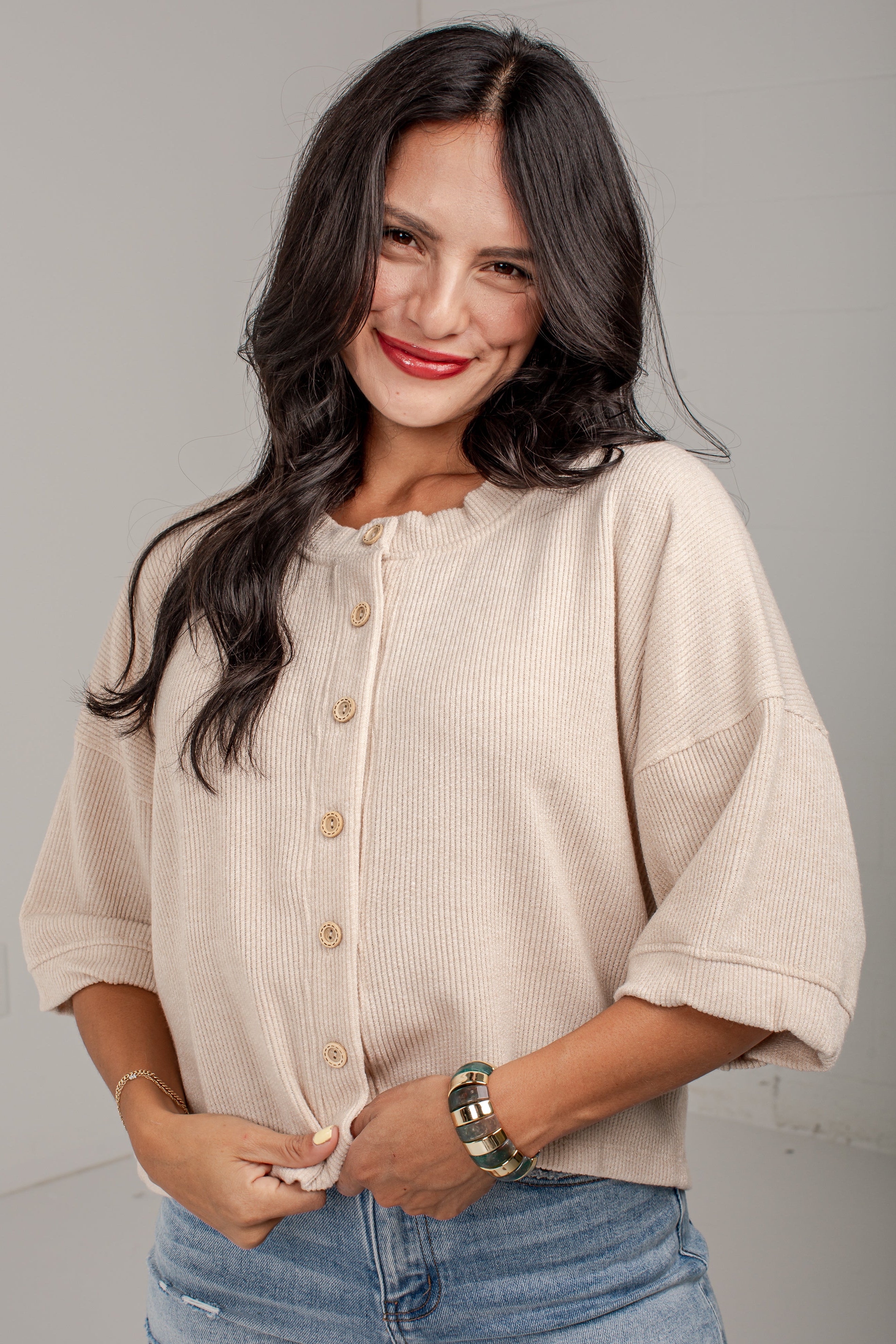 A woman with long dark hair wearing a beige short-sleeve button-front top and light blue jeans, standing against a plain light gray wall.