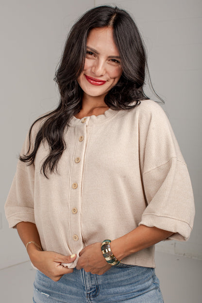 A woman with long dark hair wearing a beige short-sleeve button-front top and light blue jeans, standing against a plain light gray wall.
