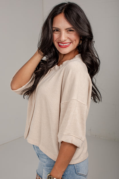 A smiling woman with long dark hair wearing a beige waffle-knit short-sleeve top and light blue jeans, posing against a light gray studio background.