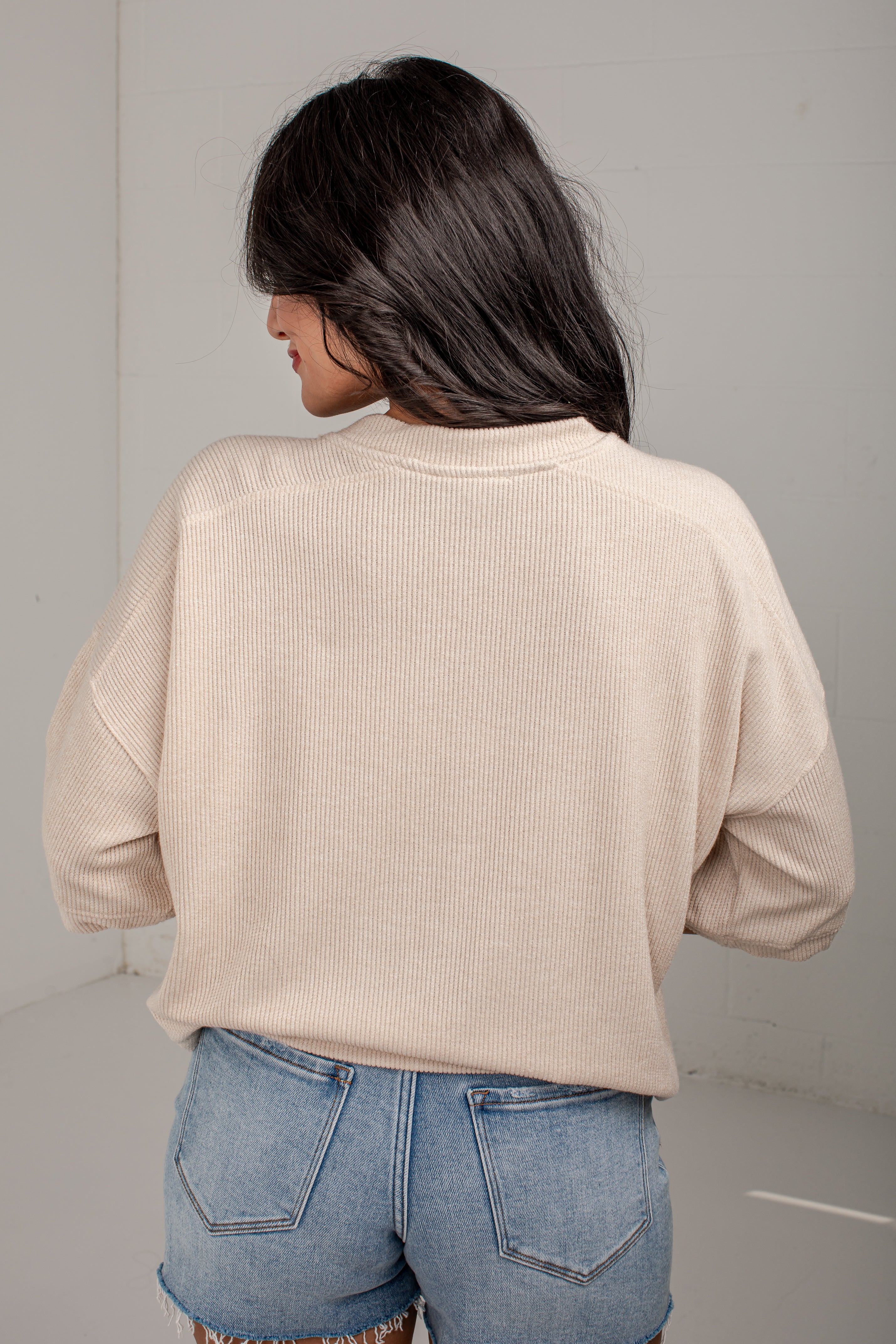 A woman shown from behind wearing a light beige ribbed knit long-sleeve top and blue denim jeans in a minimalist studio setting.