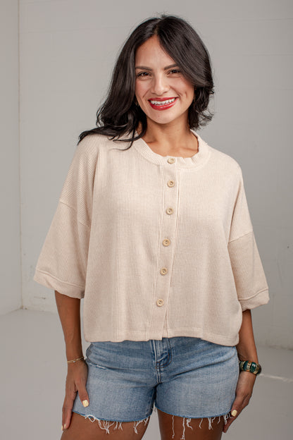 A woman wearing a cream-colored, short-sleeved button-up top with a relaxed fit and denim shorts, standing against a plain light gray wall.