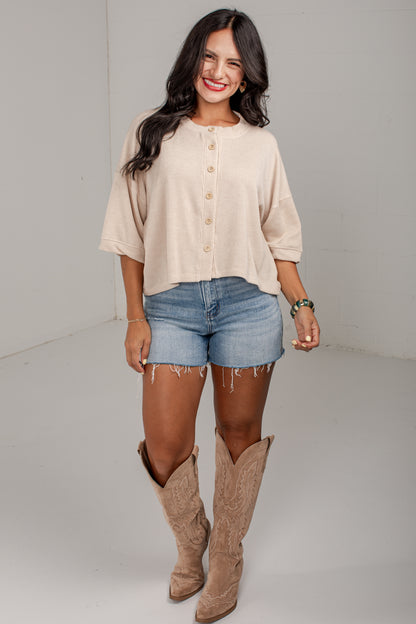 A woman modeling a beige short-sleeve button-front top, blue denim shorts, and tan cowboy boots against a plain light gray studio background.