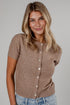 A woman with blonde hair wearing a short-sleeve knit cardigan in a warm taupe-brown color, paired with light blue jeans, against a solid gray studio background.