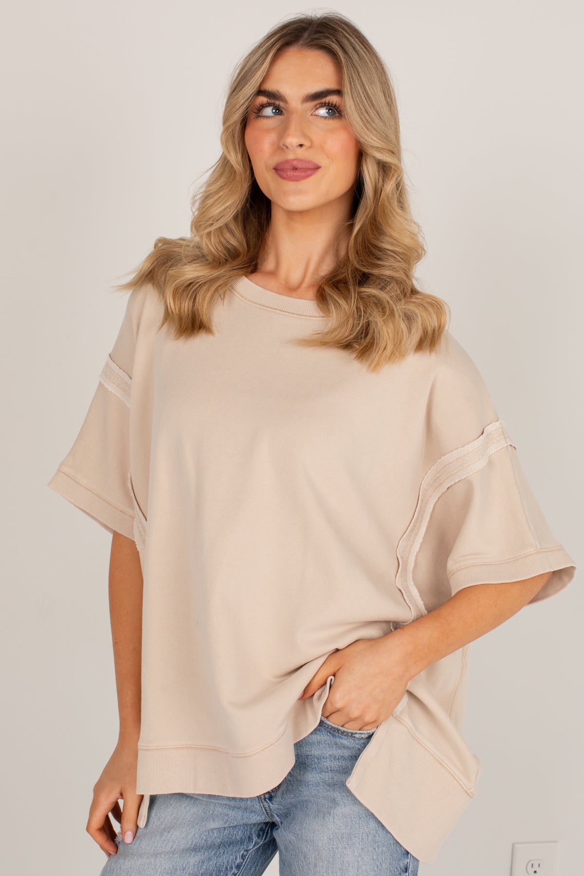 A woman wearing a loose, short-sleeve beige top with layered ruffle details on the sleeves, paired with blue jeans, against a plain light gray background.