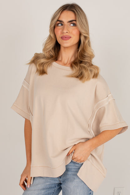 A woman wearing a loose, short-sleeve beige top with layered ruffle details on the sleeves, paired with blue jeans, against a plain light gray background.