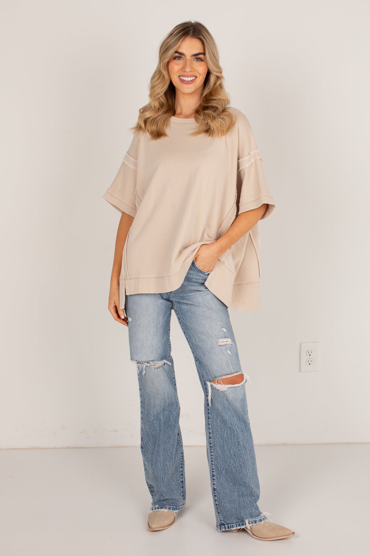 A woman modeling a beige oversized short-sleeve top with distressed details, paired with light-wash ripped jeans and beige flats, standing against a plain off-white wall.
