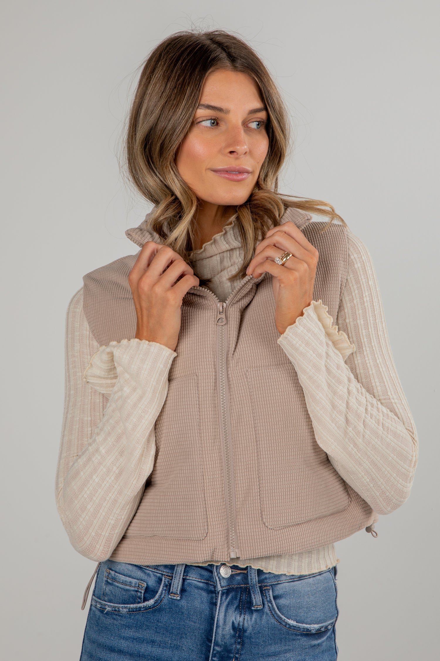 Woman wearing a beige vest over a white sweater with blue jeans on a plain background