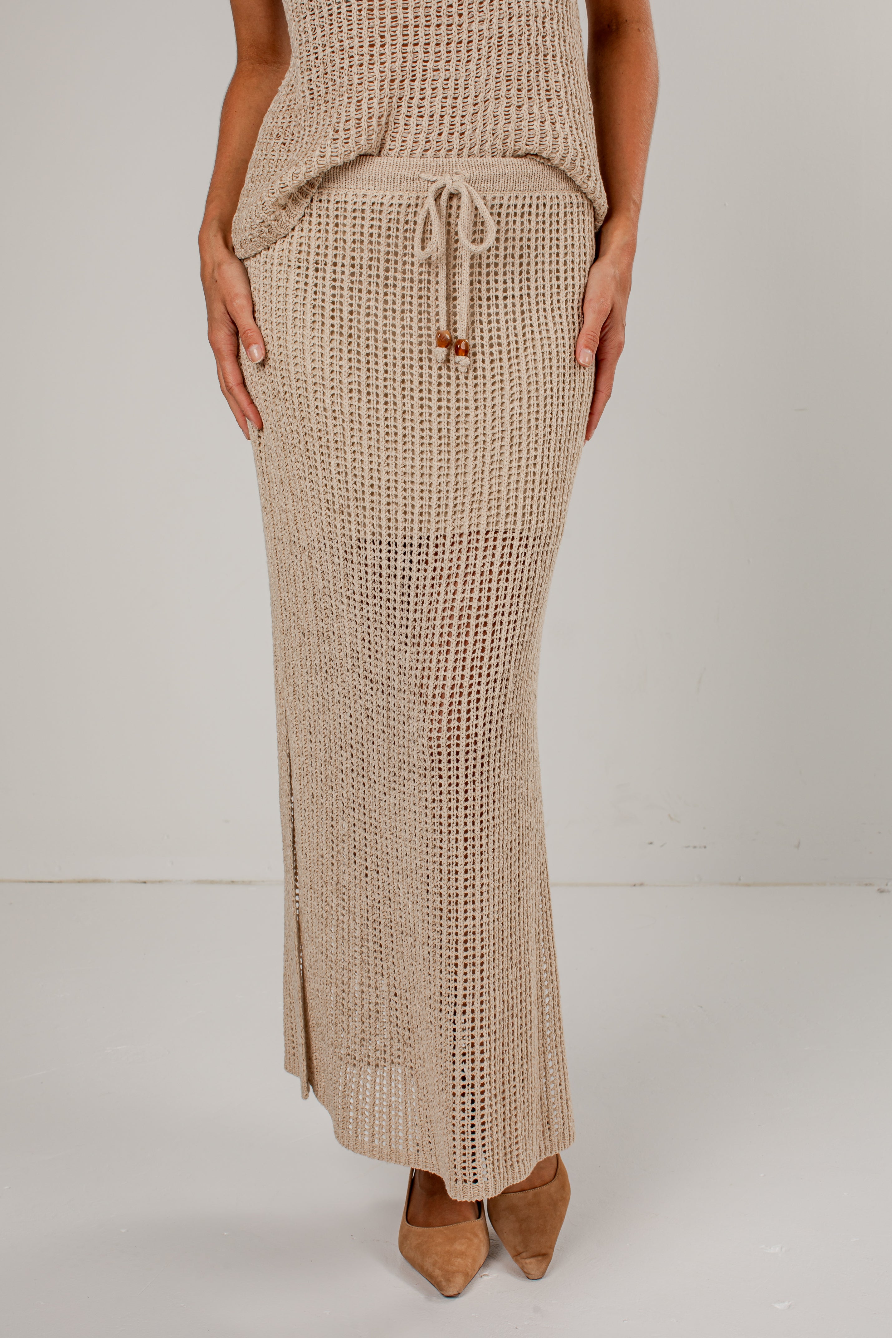 A woman wearing a beige chunky-knit maxi skirt with a drawstring waist and side slit, paired with a matching knit top and tan pointed-toe heels, against a plain light gray studio background.