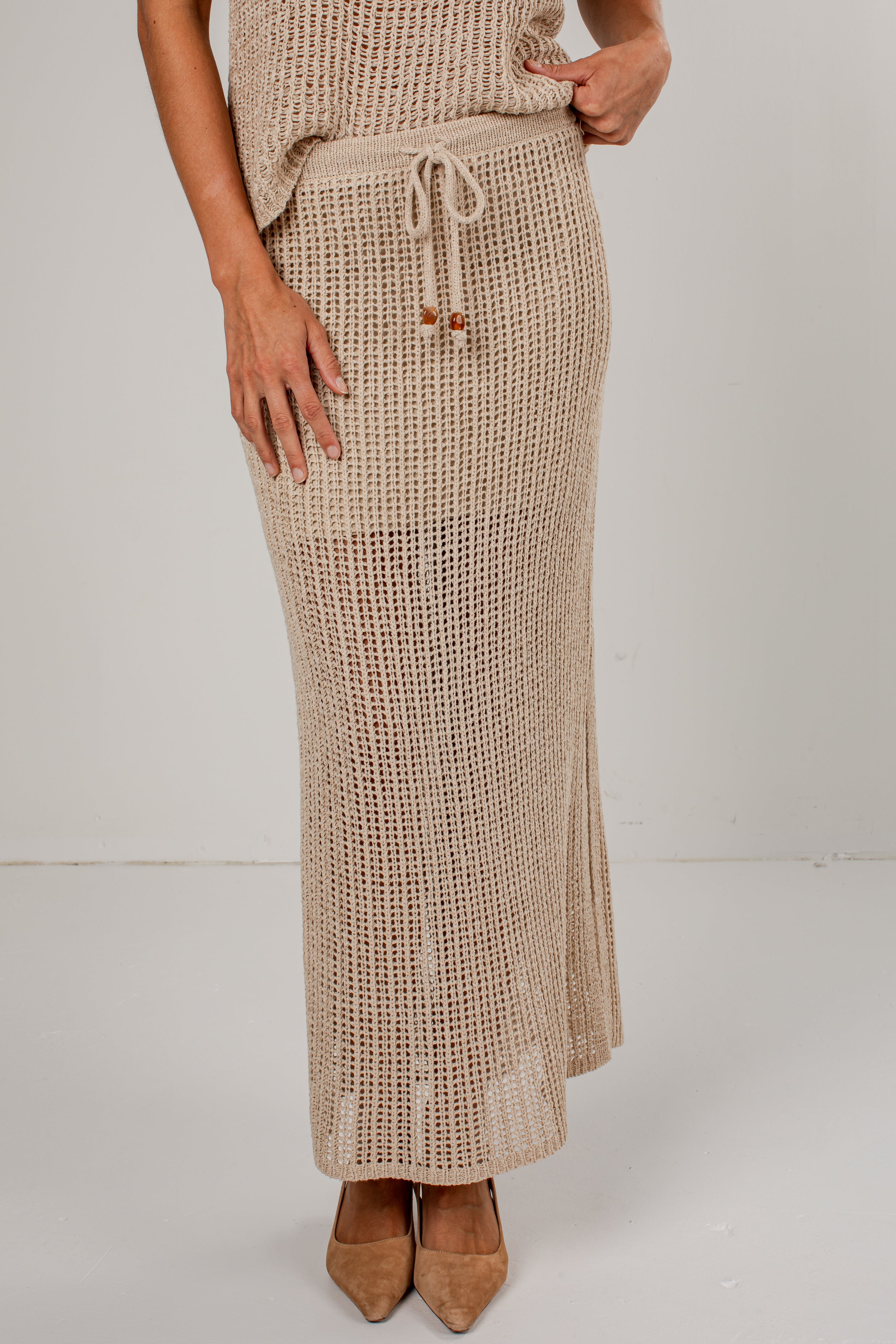 A woman wearing a beige open-knit maxi skirt with a drawstring waist, paired with a matching knit top and tan pointed-toe heels, photographed against a plain light gray studio background.