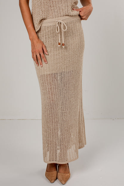 A woman wearing a beige open-knit maxi skirt with a drawstring waist, paired with a matching knit top and tan pointed-toe heels, photographed against a plain light gray studio background.