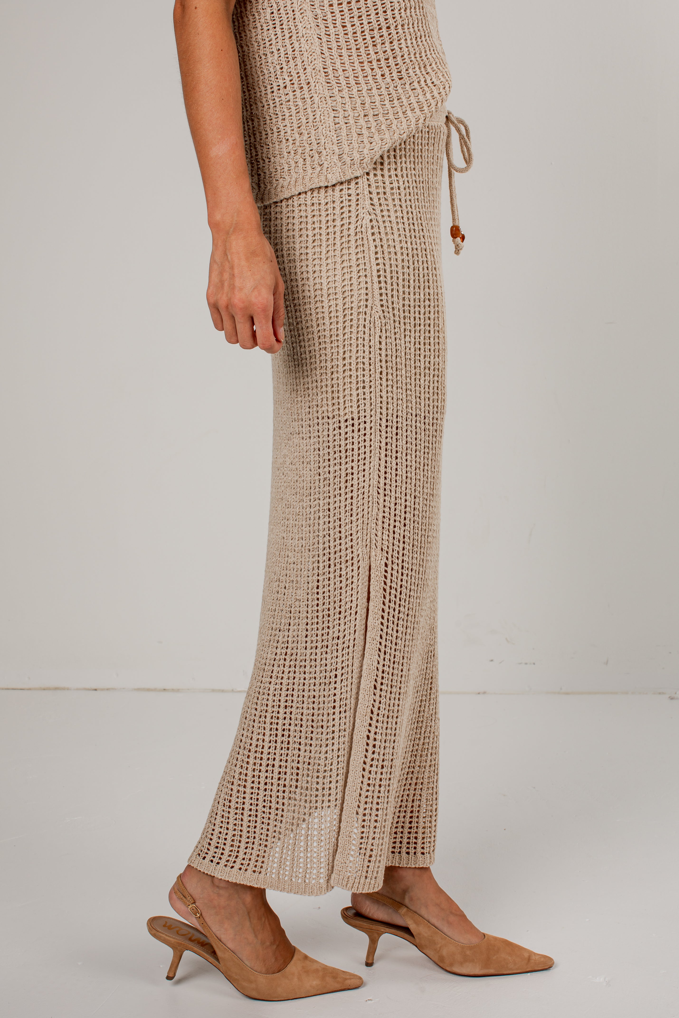 A model wearing a beige open-knit cropped vest and matching wide-leg knit pants with a side slit, paired with tan slingback heeled mules, against a plain light gray studio background.