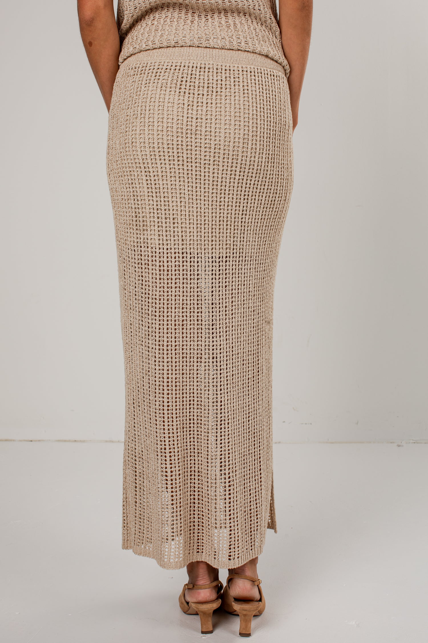 A person shown from the back wearing a long, open-knit beige maxi skirt with a matching top, paired with tan heeled sandals, against a plain light gray studio background.