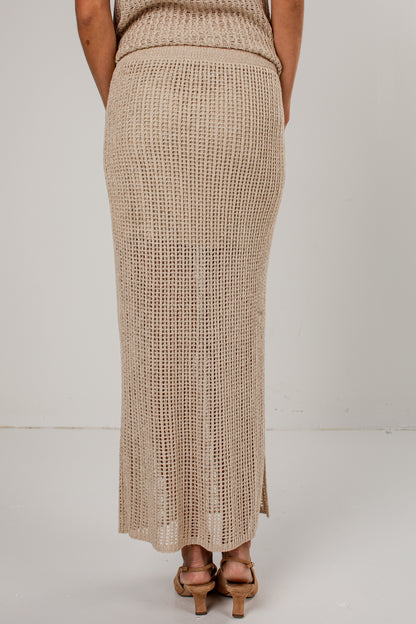 A person shown from the back wearing a long, open-knit beige maxi skirt with a matching top, paired with tan heeled sandals, against a plain light gray studio background.