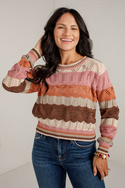 A woman smiling and wearing a long-sleeve knit sweater with horizontal stripes in pink, brown, orange, and cream, paired with blue jeans against a light gray wall.