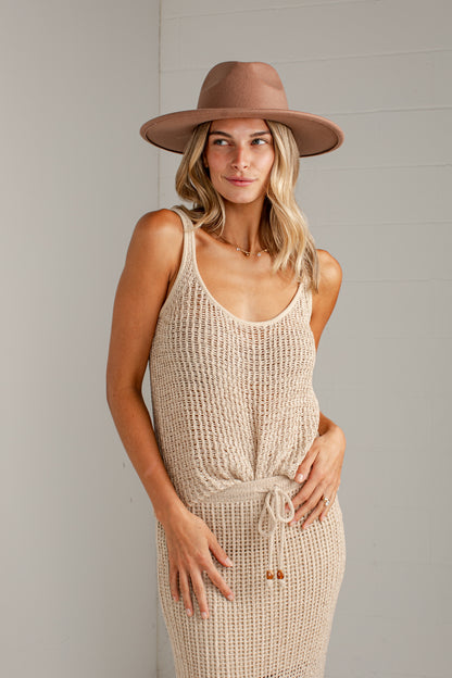 A woman wearing a beige chunky-knit sleeveless dress with a drawstring waist and a wide-brimmed taupe hat, standing against a light gray paneled wall.