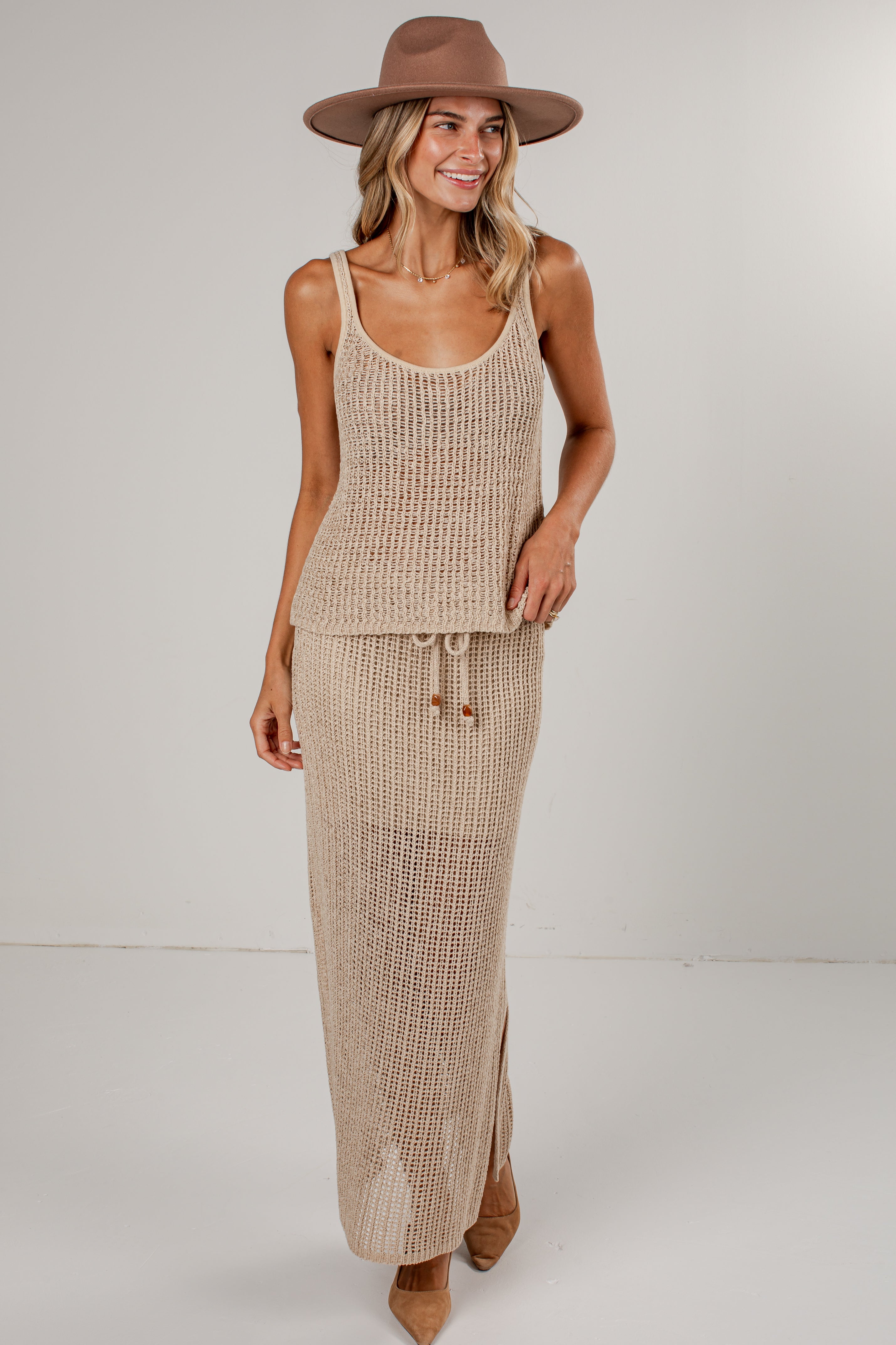 A woman modeling a beige knit two-piece set consisting of a sleeveless tank top and a long maxi skirt, styled with a brown wide-brim hat and tan pointed-toe heels against a plain light gray studio background.