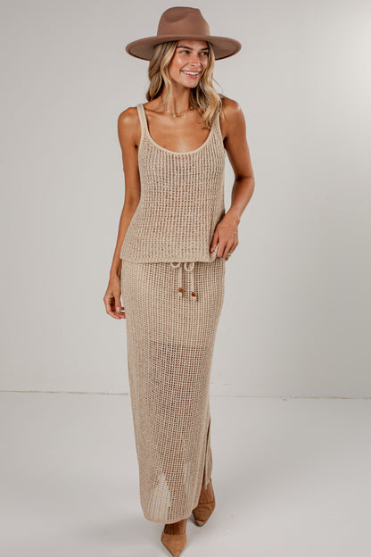 A woman modeling a beige knit two-piece set consisting of a sleeveless tank top and a long maxi skirt, styled with a brown wide-brim hat and tan pointed-toe heels against a plain light gray studio background.