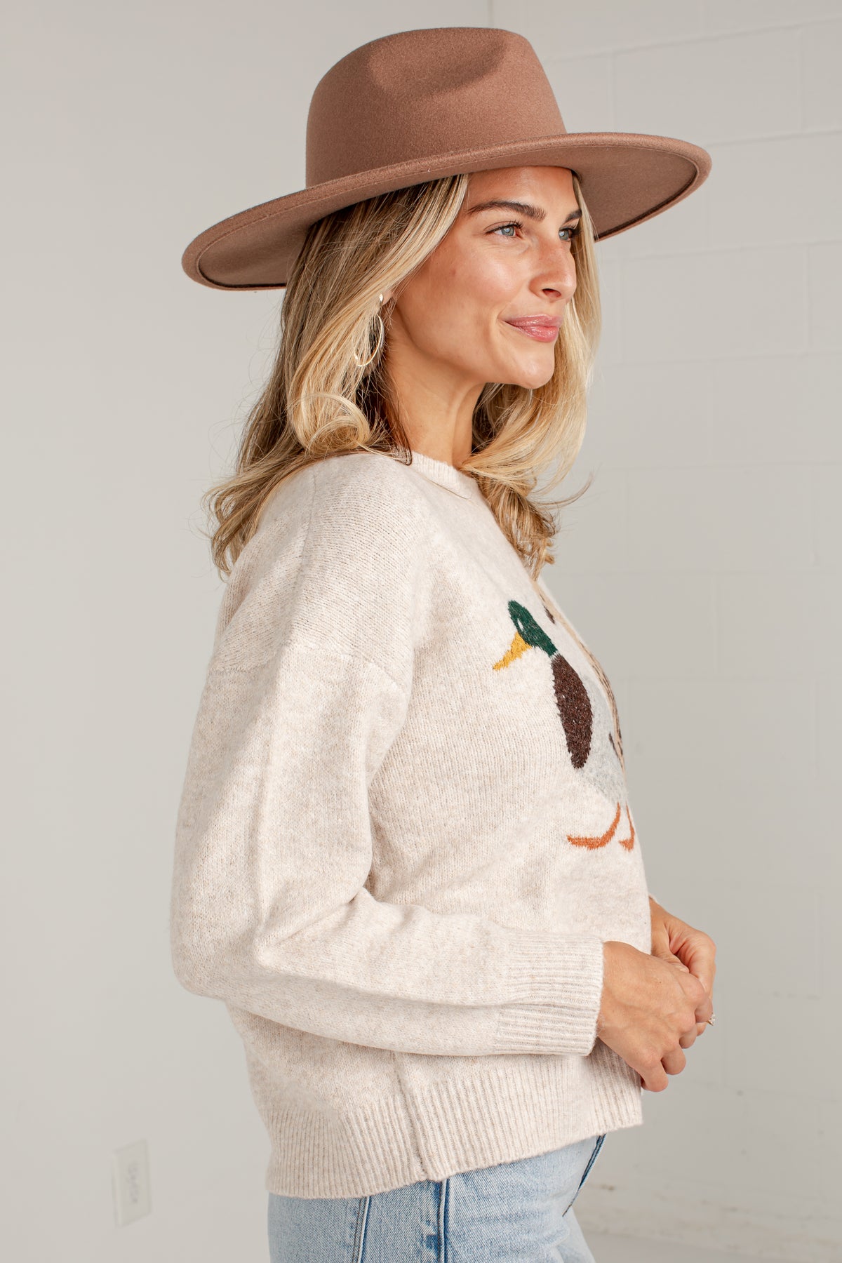 A woman wearing a cream knit sweater with a duck motif on the chest, styled with a brown wide-brim hat and light blue jeans, shown in profile against a light gray wall.