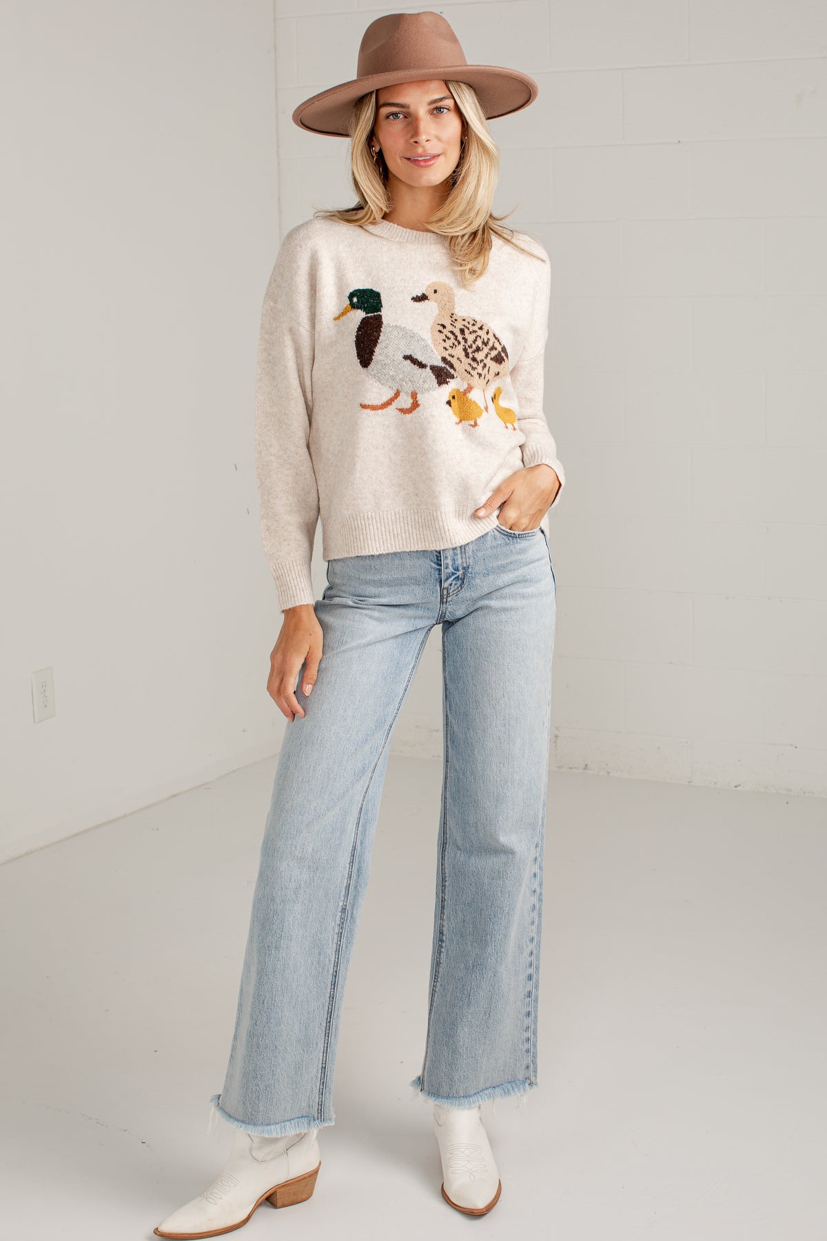 A woman modeling a cream-colored knit sweater with a detailed duck embroidery on the front, paired with light-wash wide-leg jeans, white boots, and a brown wide-brim hat in a bright studio setting.