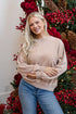 Woman in a beige sweater and blue jeans standing in front of a Christmas tree with red berries and pinecones.