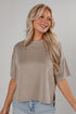 A smiling blonde woman wearing a taupe short-sleeve crop top and light blue jeans, photographed against a neutral light gray studio background.