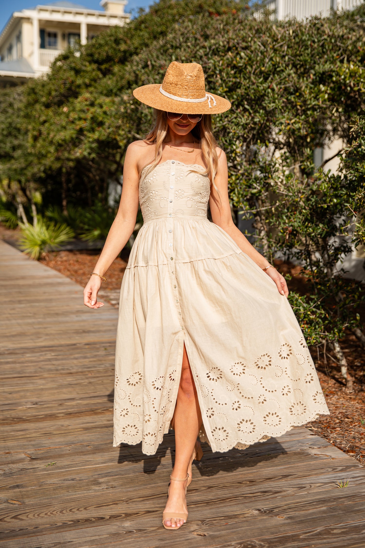 Woman in a beige dress and hat walking on a wooden path with greenery in the background