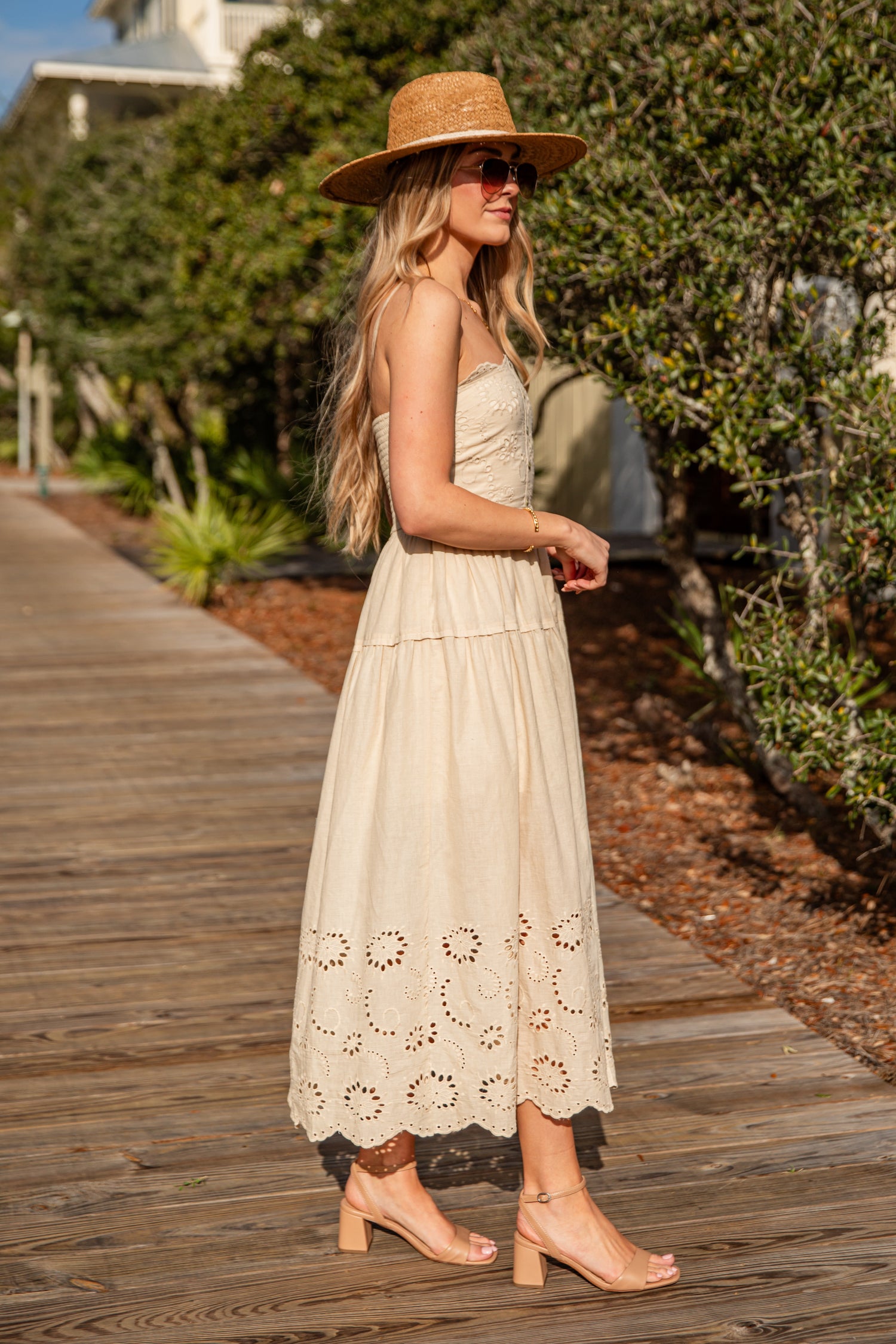 Woman in a beige dress and hat walking on a wooden path with greenery in the background
