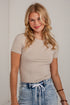 A woman with blonde hair wearing a beige short-sleeve crewneck t-shirt and light-wash blue jeans with a white drawstring, standing against a white brick wall.