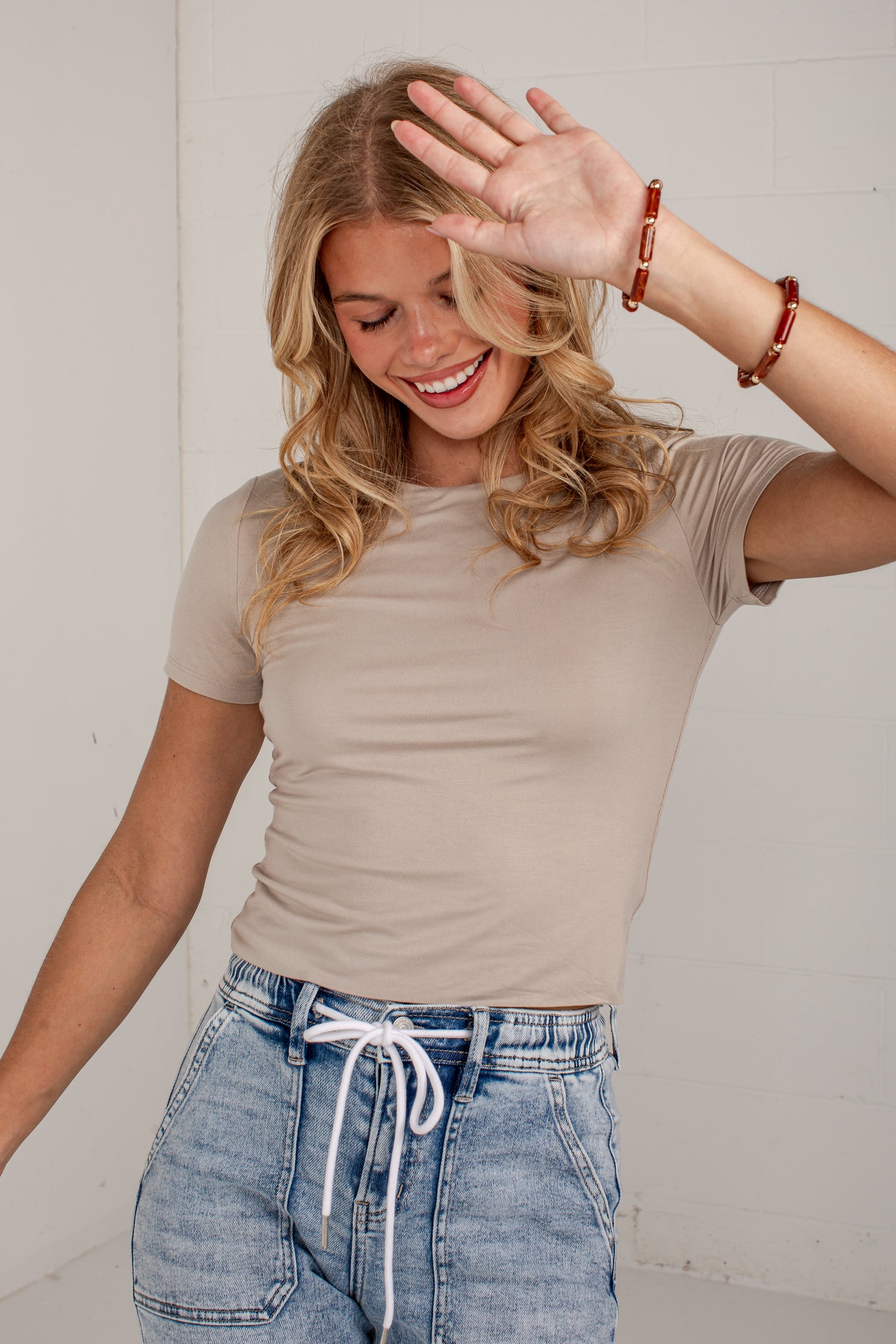 A woman wearing a beige short-sleeve t-shirt and light-wash blue jeans with a white drawstring, posing in a bright indoor setting.