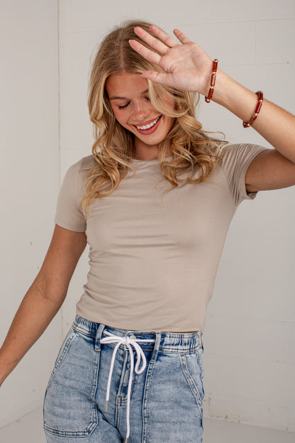 A woman wearing a beige short-sleeve t-shirt and light-wash blue jeans with a white drawstring, posing in a bright indoor setting.