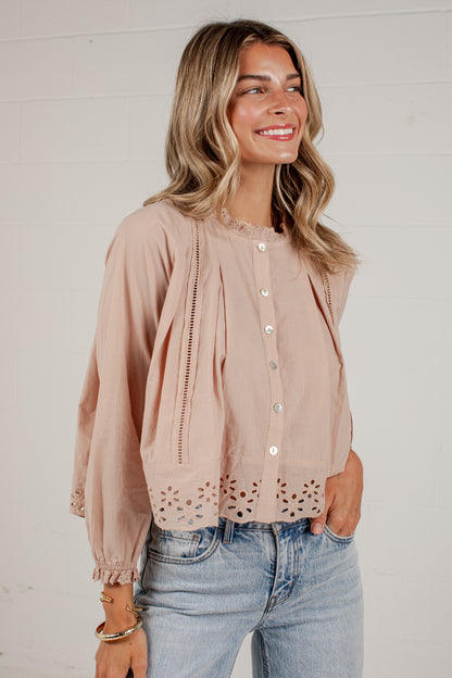 A woman wearing a light dusty pink eyelet blouse with button-front closure and gathered sleeves, paired with light-wash blue jeans, standing against a white paneled wall.